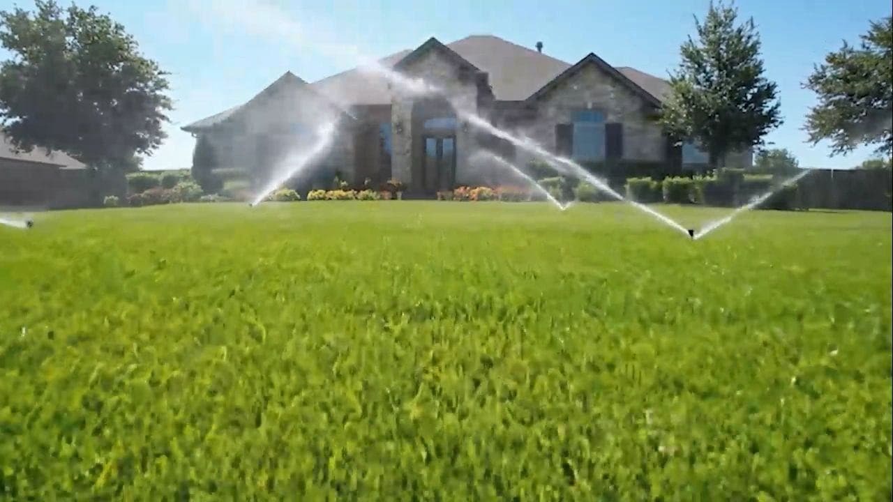 Sprinklers running on a lawn in front of a stone house.